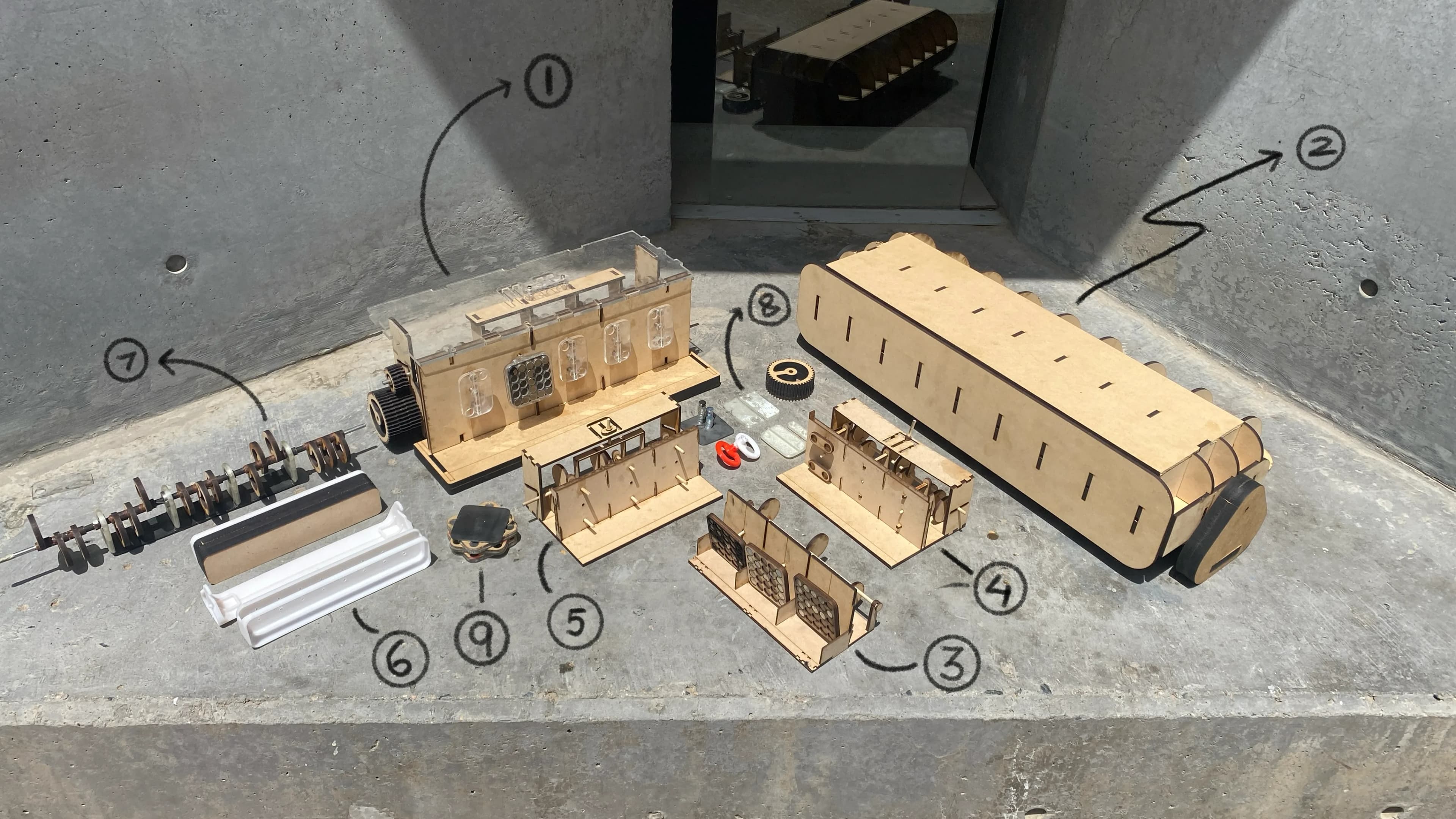 Nine build iterations arranged on a concrete surface and numbered in order: a mechanical proof of concept, a 1:1 contour scale model, three mechanism iterations culminating in a gated shifter, a 1:3 appearance model, its moving camshaft, 3D-printed housing iterations, and the final mild-steel and acrylic housing.