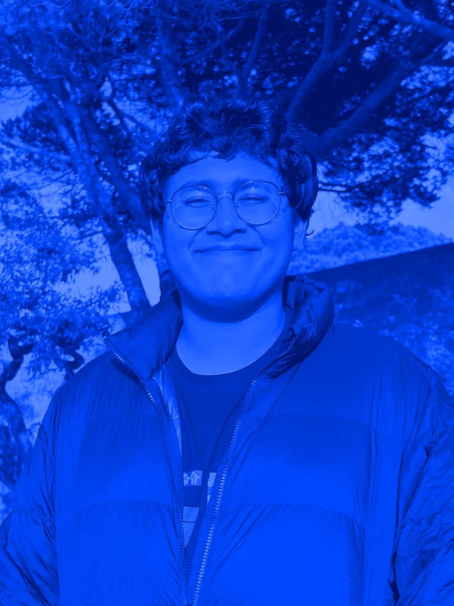 Vidush, smiling, in glasses and a dark puffer jacket, under pine trees